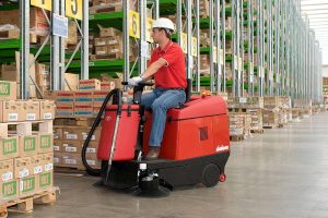 Ride-on floor scrubber cleaning warehouse aisle, illustrating equipment used when auditing a commercial cleaning contract setup
