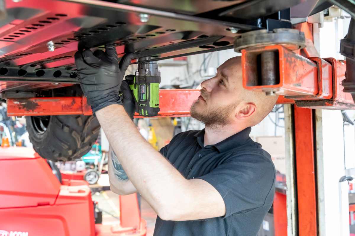 Industrial cleaning machine technician servicing the underside components of a ride-on floor scrubber dryer in a maintenance workshop